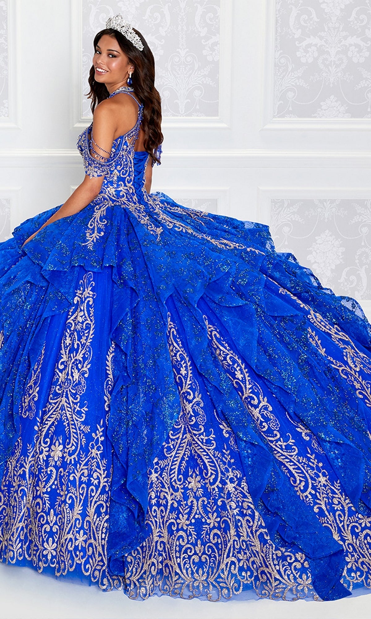 A woman wearing a tiara and the Princesa PR12274 Quinceañera Dress with Glitter, featuring gold embroidery, stands against a white patterned wall.